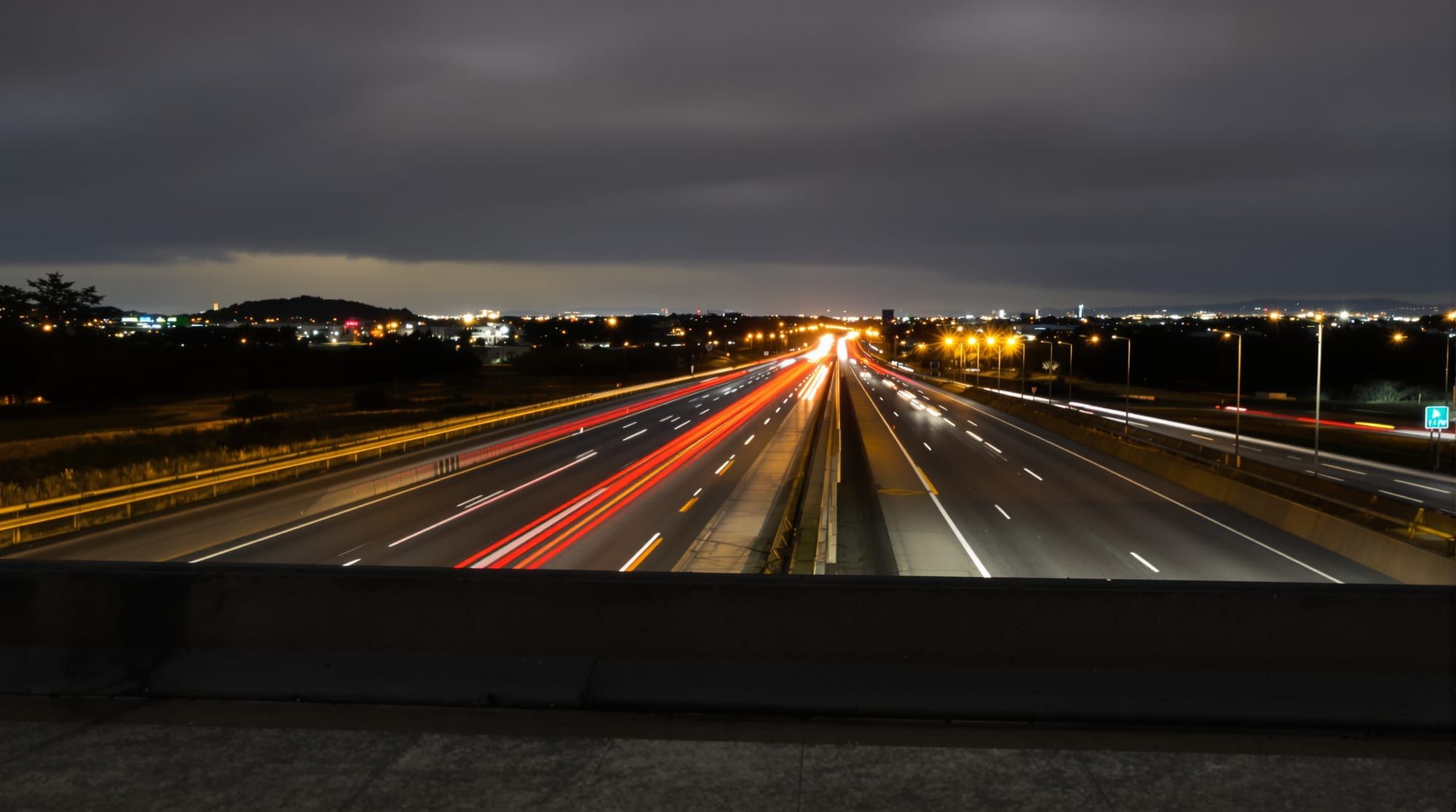 Interstate at night from an overpass, red and white light trails under a cloudy sky